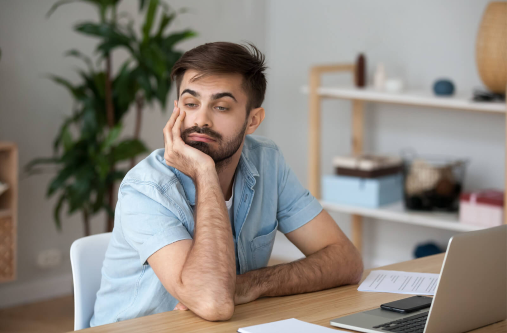 A young man sitting in front of a laptop in an office and looking away, bored, while resting his chin on his right hand.