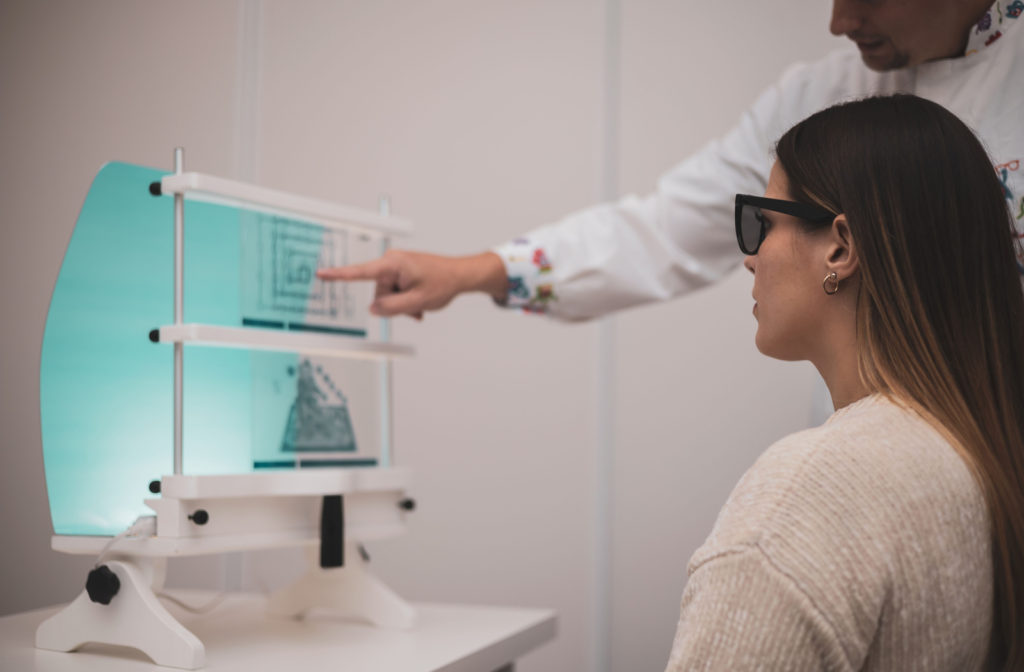 Side-view of a young woman wearing dark glasses and looking at a diagram as her eye doctor points to the middle of it.