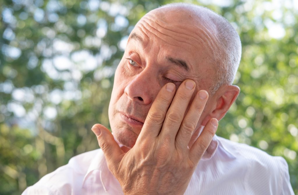 An older adult in a white shirt standing outside and rubbing their left eye due to allergies.