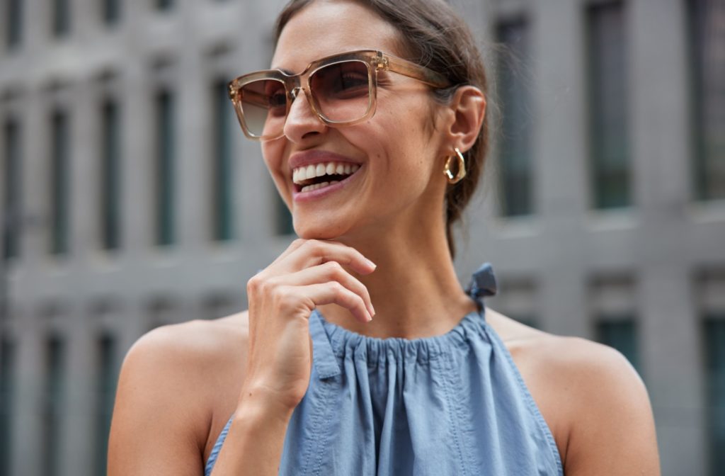 A woman wearing sunglasses and smiling with her hand to her chin.