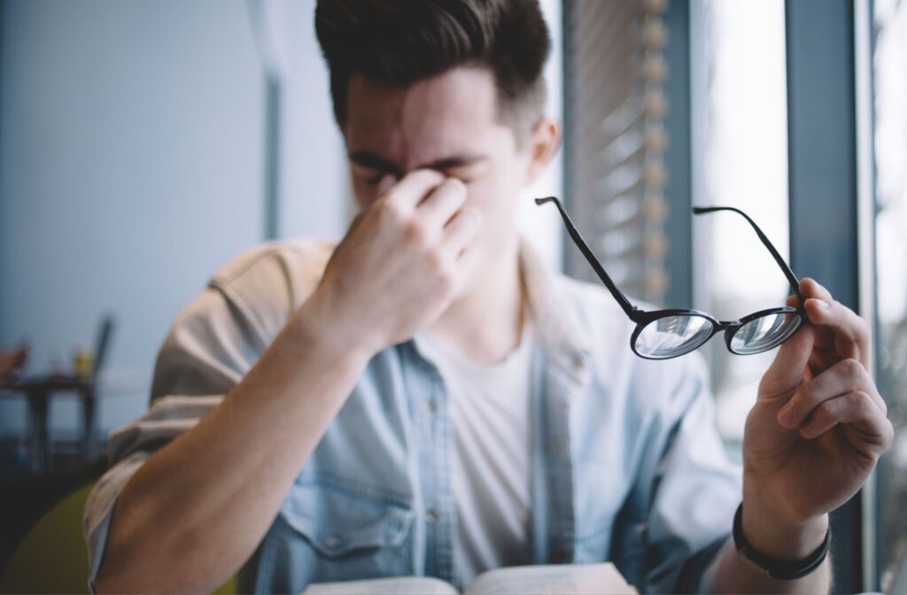 A young adult holds their glasses and rubs their tired eyes after spending some time reading.