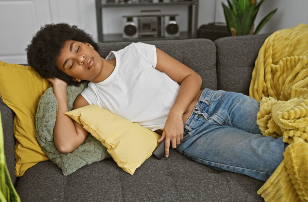 Young adult lying on their side on the couch with their head on one hand, holding a TV remote with the other.