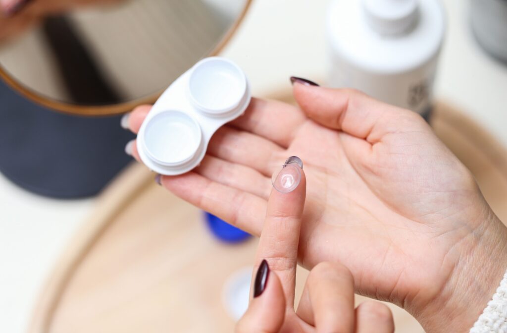 A close-up of well-manicured hands handling contact lenses—one hand holding a contact lens case, the other displaying a lens balanced on a fingertip.