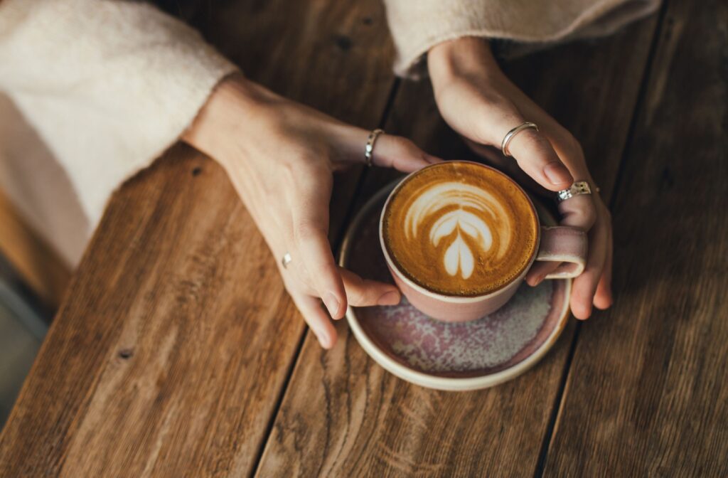 A birds-eye view of a cup of coffee sitting on a wood table with hands holding the cup
